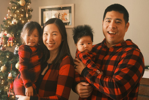 A family wearing matching red and black plaid outfits standing in front of a decorated Christmas tree, smiling warmly. The family includes a mother holding a young daughter, a father holding a baby, and a cozy holiday setting