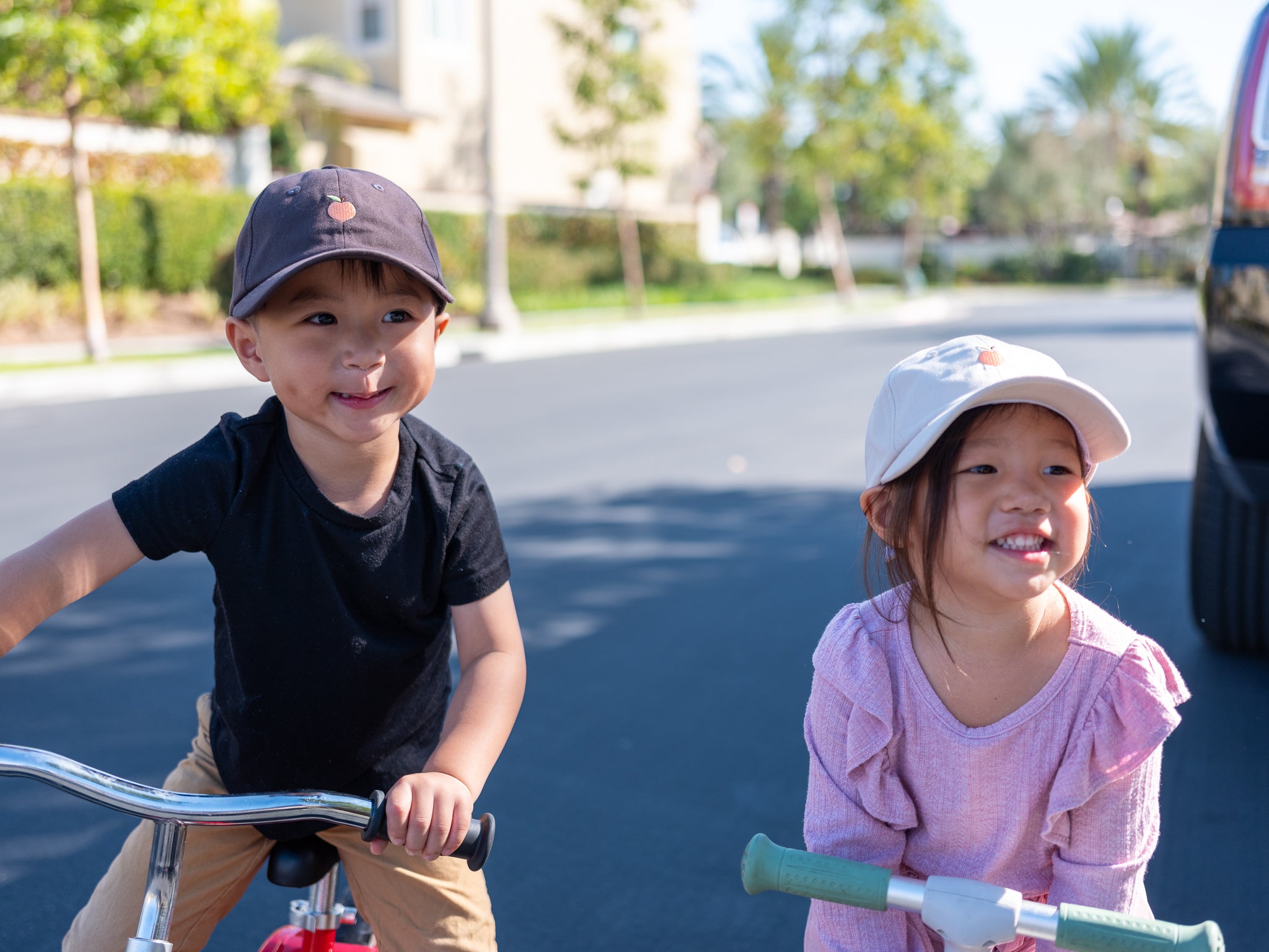 Two young children smiling while riding balance bikes outdoors on a sunny day, wearing matching hats with a peach logo, enjoying a playful moment in a suburban neighborhood